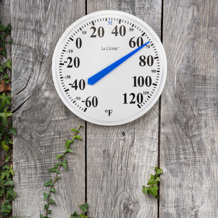 The 104-114A 13.5-inch dial thermometer with key hider with a white dial and black numbers in degrees Fahrenheit from minus 60 to 120 degrees with a blue pointer highlighting freezing at 32 degrees hanging on a weathered barn board fence with a green vine growing up the left side