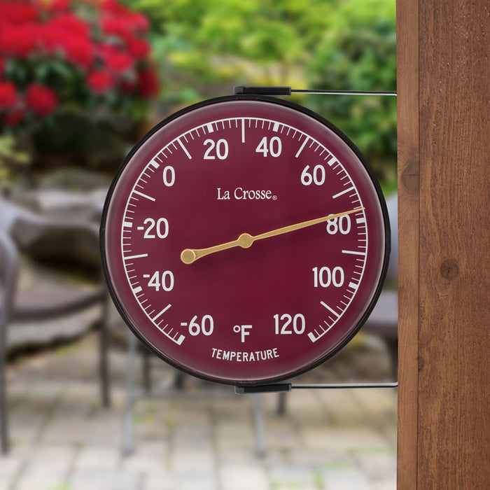 Lifestyle image of the 104-1512D 5-inch Dial Thermometer with Bracket features a wine red dial with white temperature readings in a Fahrenheit scale from -60 to 120 degrees, with an antique gold pointer shown mounted on a post in a backyard paver patio with trees and a red flowering bush in the background