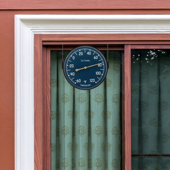 Lifestyle image of the 104-1512E 5-inch Dial Thermometer with Bracket features a navy blue dial with white temperature readings in a Fahrenheit scale from -60 to 120 degrees, with an antique gold pointer shown mounted on the frame of a window with the bracket on the top, green curtains, cedar boards, and white trim