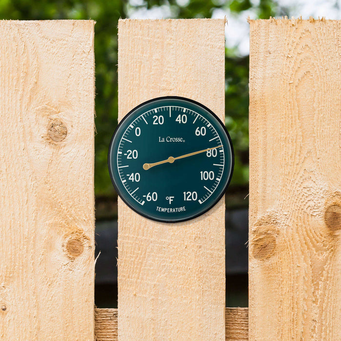 Lifestyle image of the 104-1512F 5-inch Dial Thermometer with Bracket that features an emerald green dial with white temperature readings in a Fahrenheit scale from -60 to 120 degrees, with an antique gold pointer mounted on a natural wood fence