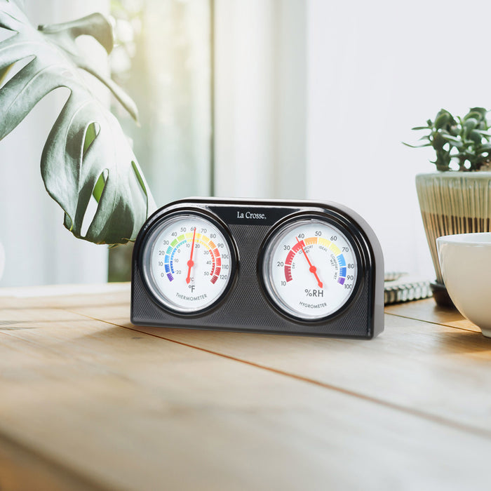 Lifestyle image of the 104-288 Analog Thermometer and Hygrometer with dual dials in a black case sitting on a wood table with a plant and bowl.