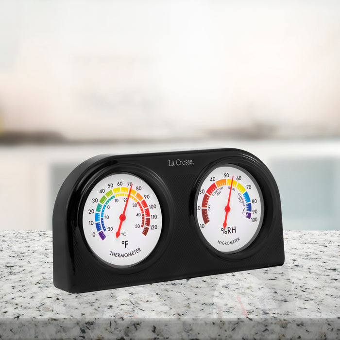 Right-facing image of the 104-288 Indoor Temperature and Humidity Gauge. Featuring dual dials with a rainbow scale, the temperature dial is on the left, reading 0-120 degrees Fahrenheit and -20-50 degrees Celsius, and the humidity dial is on the right, reading 10-100 percent RH with comfort level text. The gauge sits on a quartz countertop in grays, white, and black.