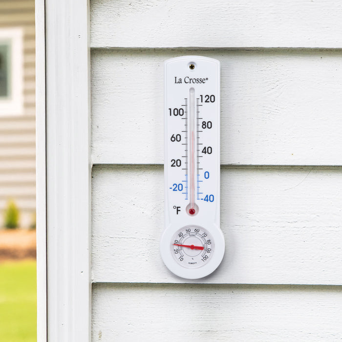 Lifestyle image of the 204-109 8.75-inch Thermometer with Hygrometer and Key Hider in a white case hanging on the outside wall of a white shed with a tan house in the background. The mercury-free capillary thermometer has a Fahrenheit temperature scale. Temperature markings below freezing appear in blue text, while those above freezing appear in black text. The Hygrometer dial reads 10-100 percent humidity, with a red pointer.