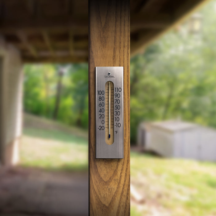 Lifestyle image of the 204-1523 9-inch Stainless Steel Capillary Thermometer shown hanging on a wood deck post with a backyard blurred out in the background. The thermometer features a brushed stainless steel finish, bold black numbers, blue capillary fill, and a La Crosse logo.