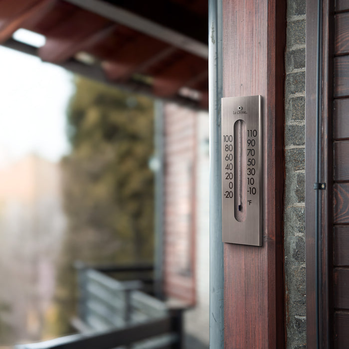Lifestyle image of the 204-1523 9-inch Stainless Steel Capillary Thermometer shown hanging on a walnut wood trim next to a window showing a view of a modern home. The thermometer features a brushed stainless steel finish, bold black numbers, blue capillary fill, and a La Crosse logo.