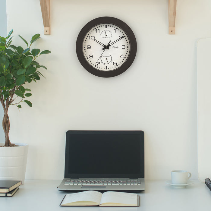 29005 10-inch Waylon indoor outdoor wall clock with temperature and humidity, linen design dial with dark brown frame features stylized cut out hands: hour and minute, and a second hand hanging above a desk with a laptop computer, book, plant and coffee cup