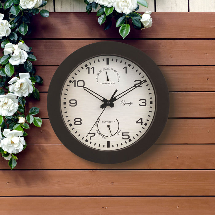 Outdoor lifestyle image of the 29005 10-inch Waylon indoor outdoor wall clock with temperature and humidity, featuring a linen design dial with a dark brown frame, stylized cut-out hour and minute hands, and a second hand hanging on a fence with flowers