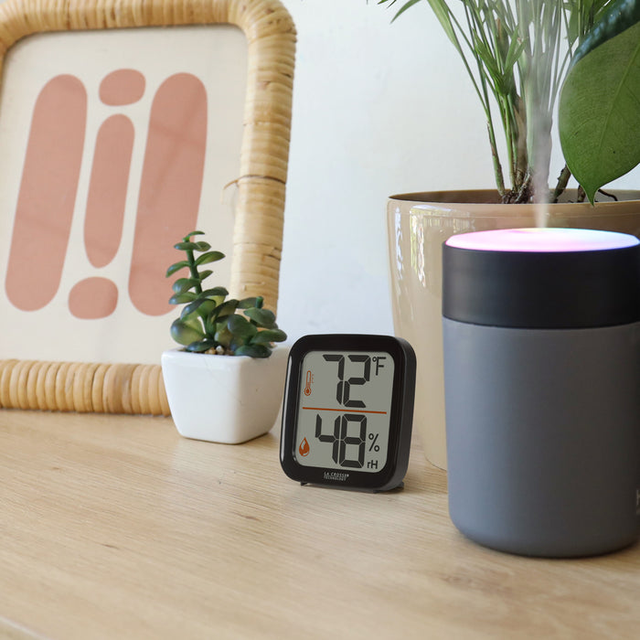 Lifestyle image of the 302-1407B Thermometer with Temperature and Humidity - Black. The thermometer is shown sitting on a light-colored wood shelf or table with plants, a picture frame, and a light-up humidifier. The thermometer features a black square case and a digital screen with large temperature and humidity readings. Next to the digits are corresponding temperature and humidity icons in orange.
