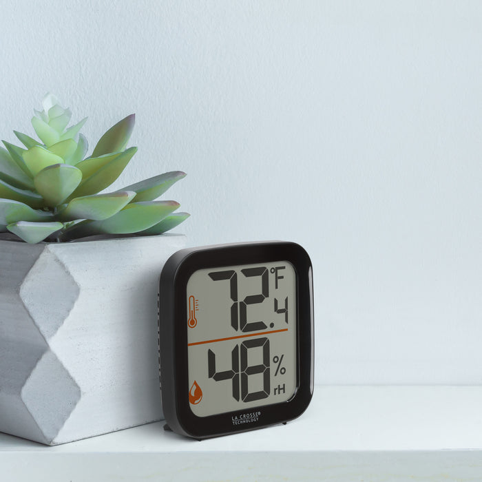 Lifestyle image of the 302-1407B Thermometer with Temperature and Humidity - Black sitting on a white shelf next to a whitewashed wood vase with succulents. The thermometer features a black square case and a digital screen with large temperature and humidity readings. Next to the digits are corresponding temperature and humidity icons in orange.