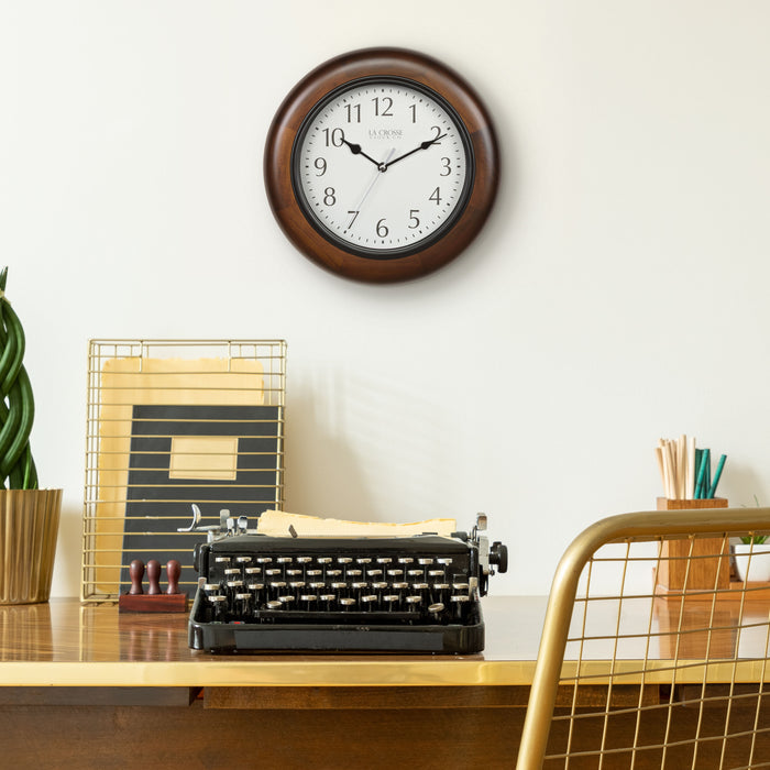 Lifestyle image of a desk with a vintage typewriter, gold mesh chair, and desk accessories with the 404-2625 Linwood wall clock hanging on a cream colored wall above the desk, the clock features a dark stain wood frame surrounding a white dial with simple black numerals and time markings, black metal hour, minute, and second hands.