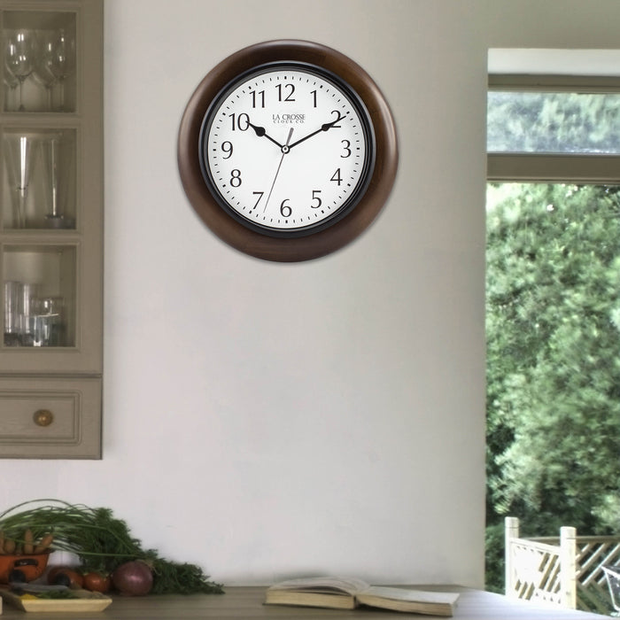 404-2625 Linwood wall clock features a dark stain wood frame surrounding a white dial with simple black numerals and time markings, black metal hour and minute hands, and a silver second hand hanging on a tan wall next to a hutch with a view of the outdoors