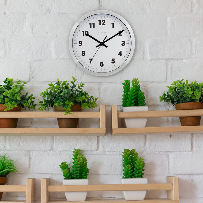 Lifestyle of the Nash 404-2626 Wall Clock with a silver metal flush mount frame surrounding a white dial with black numbers, hour, minute and second hands hanging on white brick wall above multiple open natural wood shelves with potted plants