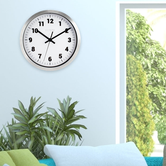 Lifestyle of the Nash 404-2626 Wall Clock with a silver metal flush mount frame surrounding a white dial with black numbers, hour, minute and second hands hanging on a blue wall in front of a couch in teal, blue, and green next to a window showing green trees