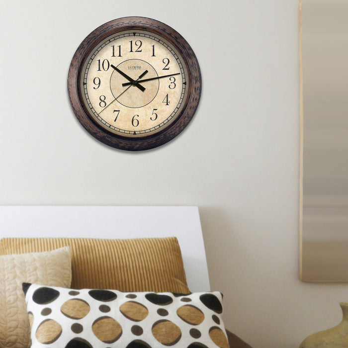 Lifestyle image of the 404-2635 14-inch Savannah Wall Clock featuring a parchment paper-inspired dial, black numbers, and a weathered bronze frame. The clock is shown hanging on a wall above a bed with gold, brown, and white bedding
