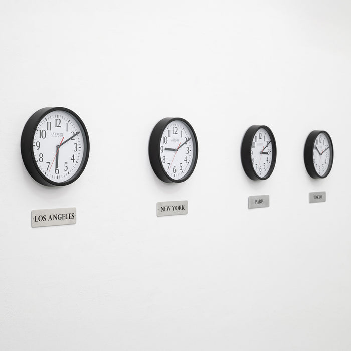 Lifestyle image of the 404-3020B-4TZ Four Pack 8-inch Basic Wall Clock with City Plaques. Four black 8-inch round wall clocks with white faces and black numerals are mounted in a horizontal row on a light gray wall. Each clock has a red second hand and displays a different time. Beneath each clock is a small rectangular plaque labeling a city: “Los Angeles,” “New York,” “Paris,” and “Tokyo,” indicating different time zones.
