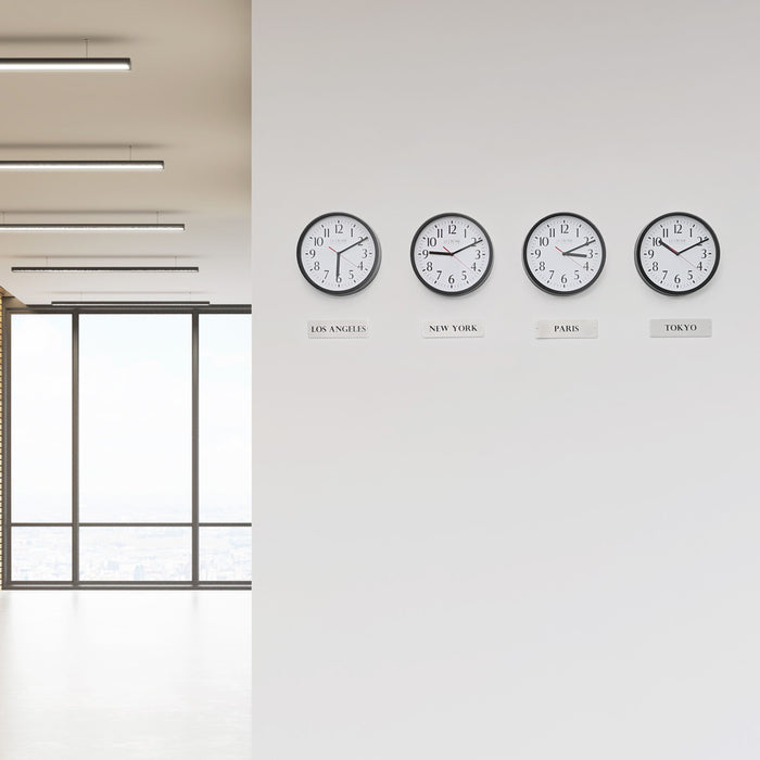 Lifestyle image of the 404-3020B-4TZ Four Pack 8-inch Basic Wall Clock with City Plaques shown hanging on a white wall in an office environment. The clocks feature black frames with white clock faces, black numbers, black hour and minute hands, and red second hands.