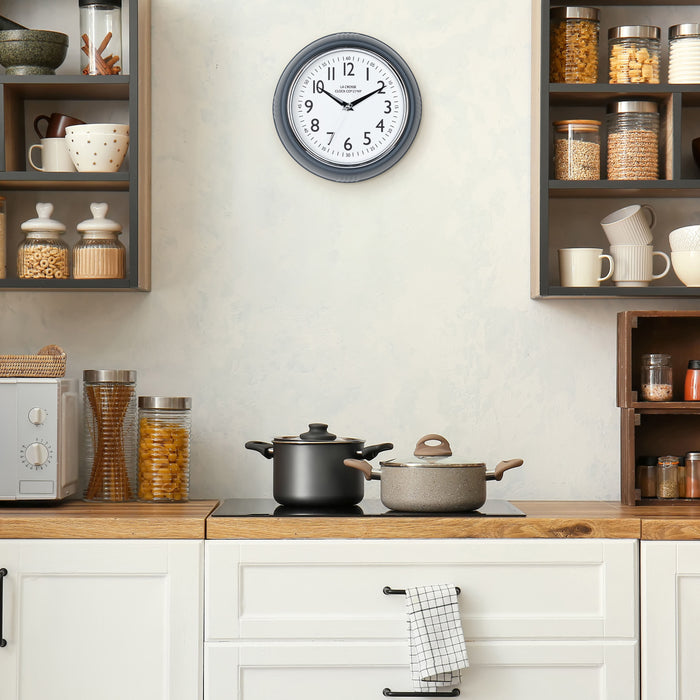 Lifestyle image of the 404-3024DS 9.5-inch Diner Clock – Dark Slate shown hanging on a cream kitchen wall above a stove top and cabinets. The clock features a retro-style dark slate frame with a white dial, chrome bezel, black numbers and minute markings, black metal hands, and a silver second hand.