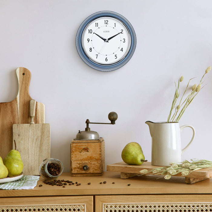 Lifestyle image of the 404-3024IB 9.5-inch Diner Clock – Indigo Blue shown hanging on a light grey wall above a wood side table with an antique coffee grinder, a white ceramic pitcher, pears, coffee, and other kitchen items. The clock features a retro-style Indigo Blue frame with a white dial, chrome bezel, black numbers and minute markings, black metal hands, and a silver second hand.