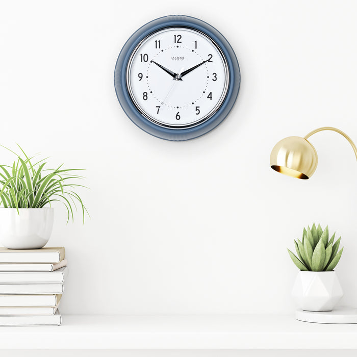 Lifestyle image of the 404-3024IB 9.5-inch Diner Clock – Indigo Blue shown hanging on a white wall above a white shelf with plants, books, and a retro lamp. The clock features a retro-style Indigo Blue frame with a white dial, chrome bezel, black numbers and minute markings, black metal hands, and a silver second hand.