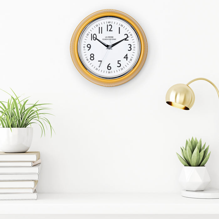 Lifestyle image of the 404-3024MY 9.5-inch Diner Clock – Mustard Yellow shown hanging on a white wall above a white shelf with plants, books, and a retro lamp. The clock features a retro-style mustard yellow frame with a white dial, chrome bezel, black numbers and minute markings, black metal hands, and a silver second hand.