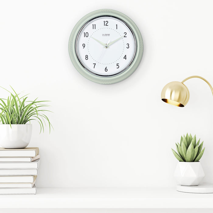 Lifestyle image of the 404-3024PH 9.5-inch Diner Wall Clock - Pistachio shown hanging on a white wall above a white shelf with books and plants. The clock features an off-white dial with black numbers, pistachio hour dots, pistachio hour and minute hands, and a silver accent second hand set within a vintage-retro pistachio green frame.
