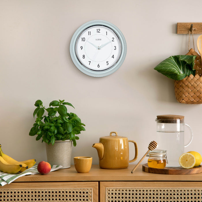 Lifestyle image of the 404-3024PH 9.5-inch Diner Wall Clock - Pistachio hanging on a tan wall above a cold or hot tea bar with a plant and fruit. The clock features an off-white dial with black numbers, pistachio hour dots, pistachio hour and minute hands, and a silver accent second hand set within a vintage-retro pistachio green frame.