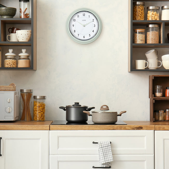 Lifestyle image of the 404-3024PH 9.5-inch Diner Wall Clock - Pistachio shown hanging on a cream kitchen wall above a stove top and cabinets. The clock features an off-white dial with black numbers, pistachio hour dots, pistachio hour and minute hands, and a silver accent second hand set within a vintage-retro pistachio green frame.