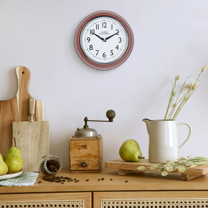 Lifestyle image of the 404-3024TC 9.5-inch Diner Clock – Terracotta shown hanging on a light grey wall above a wood side table with an antique coffee grinder, a white ceramic pitcher, pears, coffee, and other kitchen items. The clock features a retro-style terracotta frame with a white dial, chrome bezel, black numbers and minute markings, black metal hands, and a silver second hand.