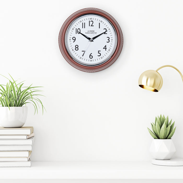Lifestyle image of the 404-3024TC 9.5-inch Diner Clock – Terracotta shown hanging on a white wall above a white shelf with plants, books, and a retro lamp. The clock features a retro-style terracotta frame with a white dial, chrome bezel, black numbers and minute markings, black metal hands, and a silver second hand.