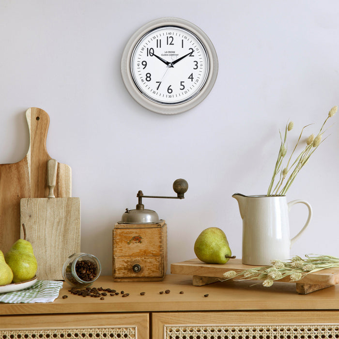 Lifestyle image of the 404-3024TP 9.5-inch Diner Clock – Taupe shown hanging on a light grey wall above a wood side table with an antique coffee grinder, a white ceramic pitcher, pears, coffee, and other kitchen items. The clock features a retro-style taupe frame with a white dial, chrome bezel, black numbers and minute markings, black metal hands, and a silver second hand.