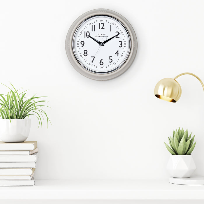 Lifestyle image of the 404-3024TP 9.5-inch Diner Clock – Taupe shown hanging on a white wall above a white shelf with plants, books, and a retro lamp. The clock features a retro-style taupe frame with a white dial, chrome bezel, black numbers and minute markings, black metal hands, and a silver second hand.