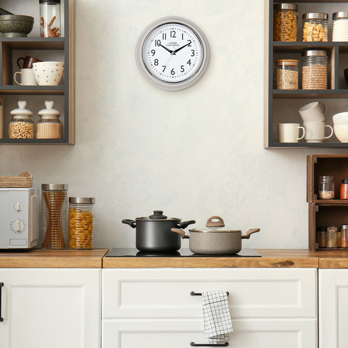 Lifestyle image of the 404-3024TP 9.5-inch Diner Clock – Taupe shown hanging on a cream kitchen wall above a stove top and cabinets. The clock features a retro-style taupe frame with a white dial, chrome bezel, black numbers and minute markings, black metal hands, and a silver second hand.
