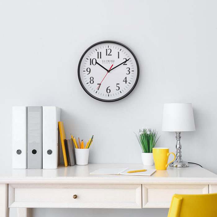 Lifestyle image of the 404-3025B 10-inch Basic Decor Wall Clock with a Black frame, white dial with black numbers, hour, and minute hands, and a red second hand hanging on a white wall above a white desk with a lamp, notebook, pencil, desk accessories, a yellow coffee mug, and a yellow chair
