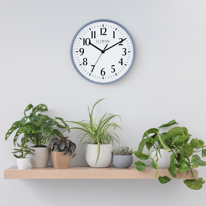 Lifestyle image of the 404-3025GB 10-inch Basic Wall Clock - Gray Blue  hanging on a light grey wall above a pine shelf with multiple plants. The clock features a white dial, black numbers and markings, and hour, minute, and second hands, surrounded by a gray blue flush-mount frame.