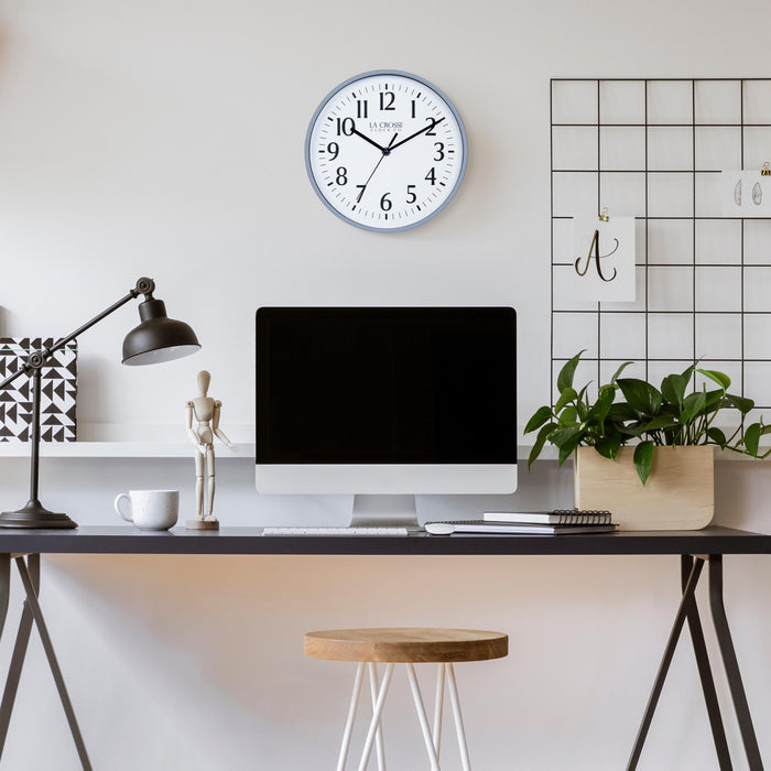 Lifestyle image of the 404-3025GB 10-inch Basic Wall Clock – Gray Blue, hanging on a light tan wall above a black desk. The desk has a computer, notebooks, a plant, a coffee mug, a black desk lamp, and a posable wood model. The clock features a white dial, black numbers, markings, hour, minute, and second hands surrounded by a gray blue flush-mount frame.