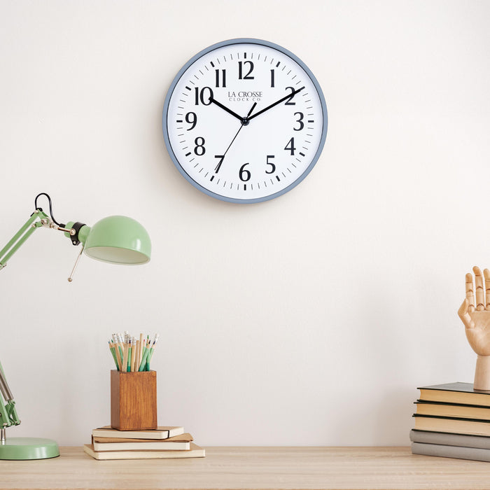 Lifestyle image of the 404-3025GB 10-inch Basic Wall Clock – Gray Blue, hanging on a light tan wall above a light wood desk with a green desk lamp, a wood pencil holder on top of a stack of notebooks, and a wood modeling hand sitting on top of a pile of books. The clock features a white dial, black numbers, markings, hour, minute, and second hands surrounded by a gray blue flush-mount frame.