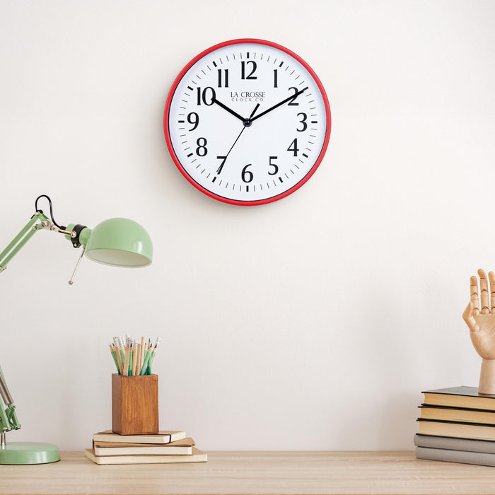 Lifestyle image of the 404-3025R 10-inch Basic Wall Clock – Red, hanging on a light tan wall above a light wood desk with a green desk lamp, a wood pencil holder on top of a stack of notebooks, and a wood modeling hand sitting on top of a pile of books. The clock features a white dial, black numbers, markings, hour, minute, and second hands surrounded by a red flush-mount frame.