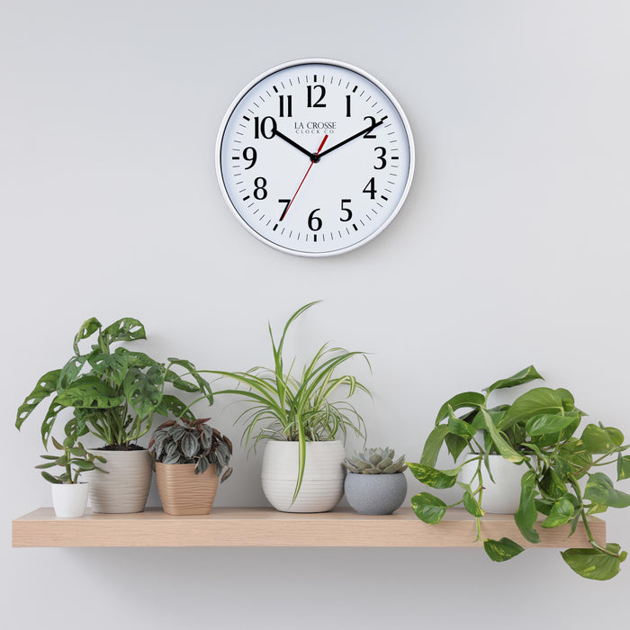 Lifestyle image of the 404-3025S 10-inch Basic Wall Clock - Silver hanging on a light grey wall above a pine shelf with multiple plants. The clock features a white dial, black numbers and markings, hour, and minute hands, and a red second hand, surrounded by a silver flush-mount frame.