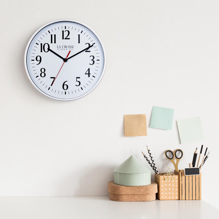 Lifestyle image of the 404-3025S 10-inch Basic Wall Clock - Silver hanging on a white wall above a white desk with cork, wood, and pastel desk accessories. The clock features a white dial, black numbers and markings, and hour, and minute hands, and a red second hand, surrounded by a silver flush-mount frame.