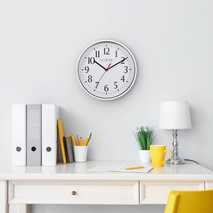 Lifestyle image of the 404-3025W 10-inch Basic Decor Wall Clock with a White frame, white dial with black numbers, hour, and minute hands, and a red second hand hanging on a white wall above a white desk with a lamp, notebook, pencil and desk accessories