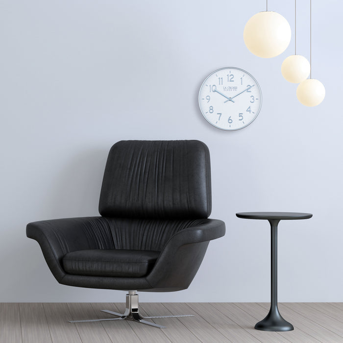 404-3027B Tovi wall clock with a white dial and silver Arabic numbers surrounded by a flush mount silver frame with matching silver hour and minute hands hanging on a neutral wall in a modern setting with a black leather chair three round globes light fixture and a black side table
