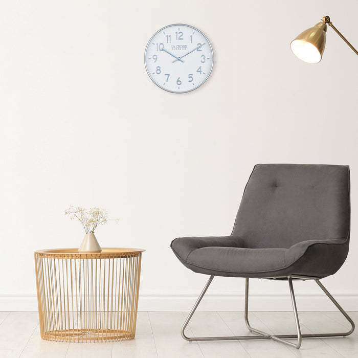 Retro decor room with the 404-3027B Tovi wall clock with a white dial and silver Arabic numbers surrounded by a flush mount silver frame with matching silver hour and minute hands hanging on a white wall with a dark gray retro chair and gold metal open cage side table with a vase