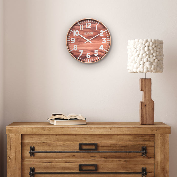 Lifestyle image of the 10.8-inch Atwood Wall Clock hanging above a rustic wood dresser with a wood lamp. The clock features a copper finish frame, a wood design dial, white numbers, and white metal hour and minute hands.