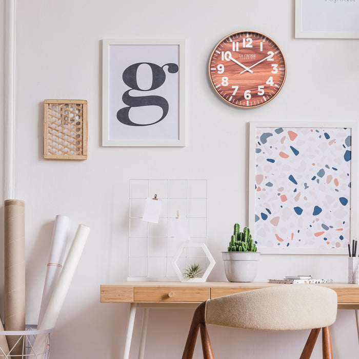 Lifestyle image of the 10.8-inch Atwood Wall Clock hanging on a wall in a home office setting with a neutral desk, fun wall art, and desk accessories. The clock features a copper finish frame, a wood design dial, white numbers, and white metal hour and minute hands.