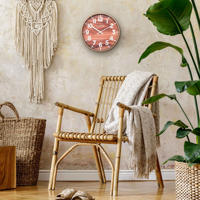 Lifestyle image of the 10.8-inch Atwood Wall Clock hanging on a tan textured wall above a wicker chair with Bohemian style decor. The clock features a copper finish frame, a wood design dial, white numbers, and white metal hour and minute hands.