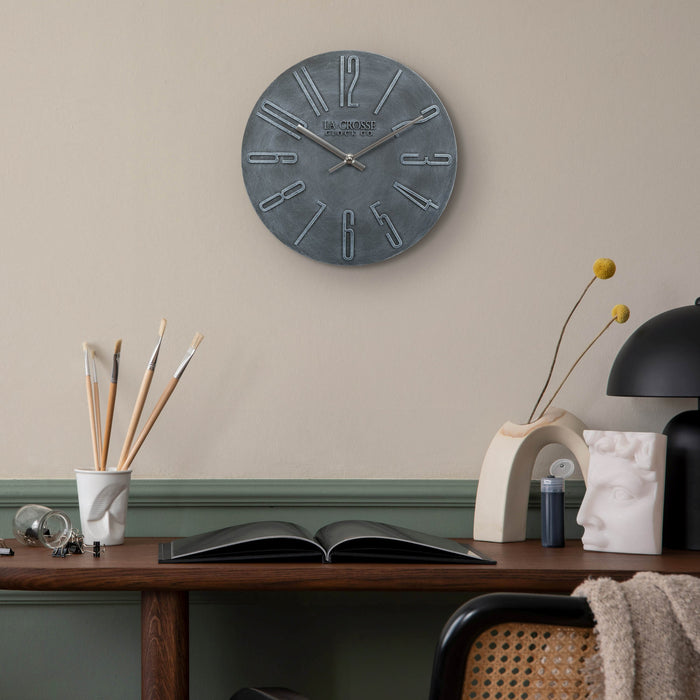 404-3028B Cormac 11 inch open face molded dark gray wall clock with numbers in relief and silver metal hour and minute hands hanging on a tan wall with green wainscotting over a dark wood desk and transitional decorative style accents