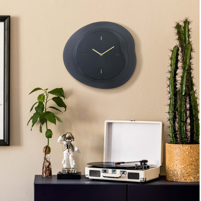 Lifestyle image of the 404-3039B 15.2-inch Drift Wall Clock – Black, shown hanging on a tan wall above a black side table with a vintage turntable, cactus, a plant, and a statue of an astronaut. The clock features an open face multi-layered design with drifting contours, metal hands, and raised number markings in a brass finish.