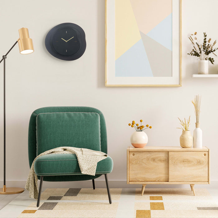 Lifestyle image of the 404-3039B 15.2-inch Drift Wall Clock – Black, shown hanging on a tan wall above an emerald green chenille chair, retro brass floor lamp, and light wood side cabinet. The clock features an open face multi-layered design with drifting contours, metal hands, and raised number markings in a brass finish.
