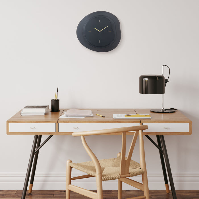 Lifestyle image of the 404-3039B 15.2-inch Drift Wall Clock – Black, shown hanging on a light wall above a modern black and wood desk. The clock features an open face multi-layered design with drifting contours, metal hands, and raised number markings in a brass finish.