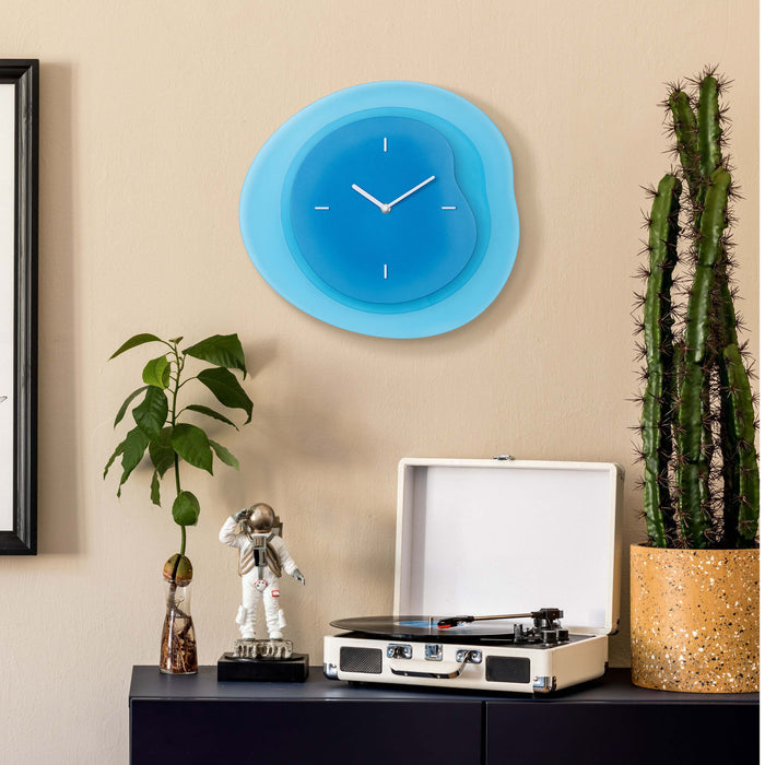 Lifestyle image of the 404-3039BL 15.2-inch Drift Wall Clock – Blue, shown hanging on a tan wall above a black side table with a vintage turntable, cactus, a plant, and a statue of an astronaut. The clock features an open face multi-layered design with drifting contours, metal hands, and raised number markings in white.